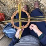 A women holding a progressing willow tension platter on her lap, as she weaves more brightly coloured rods in to fill the circle.