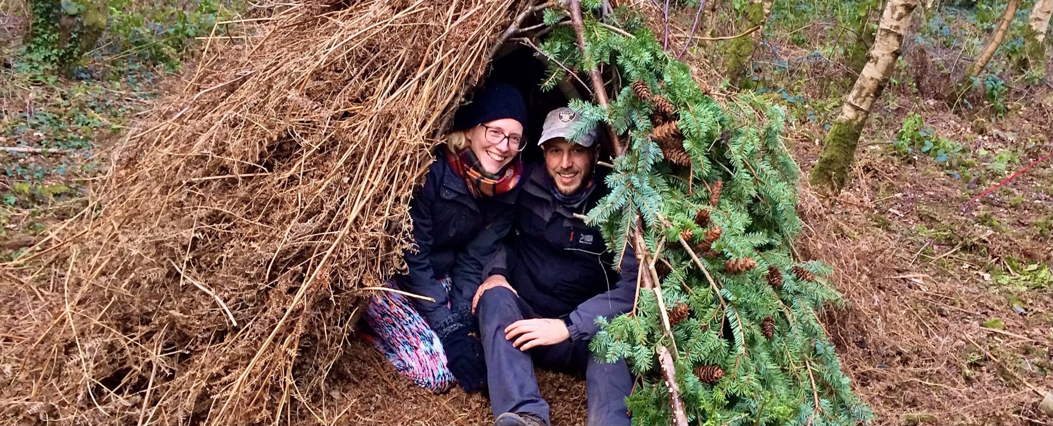 A man and a woman smiling happily in a natural debris shelter built on a weekend bushcraft course
