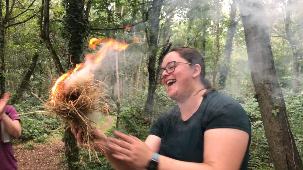 A flaming tinder bundle, and a very joyful woman as she looks on at her achievements of making friction fire.