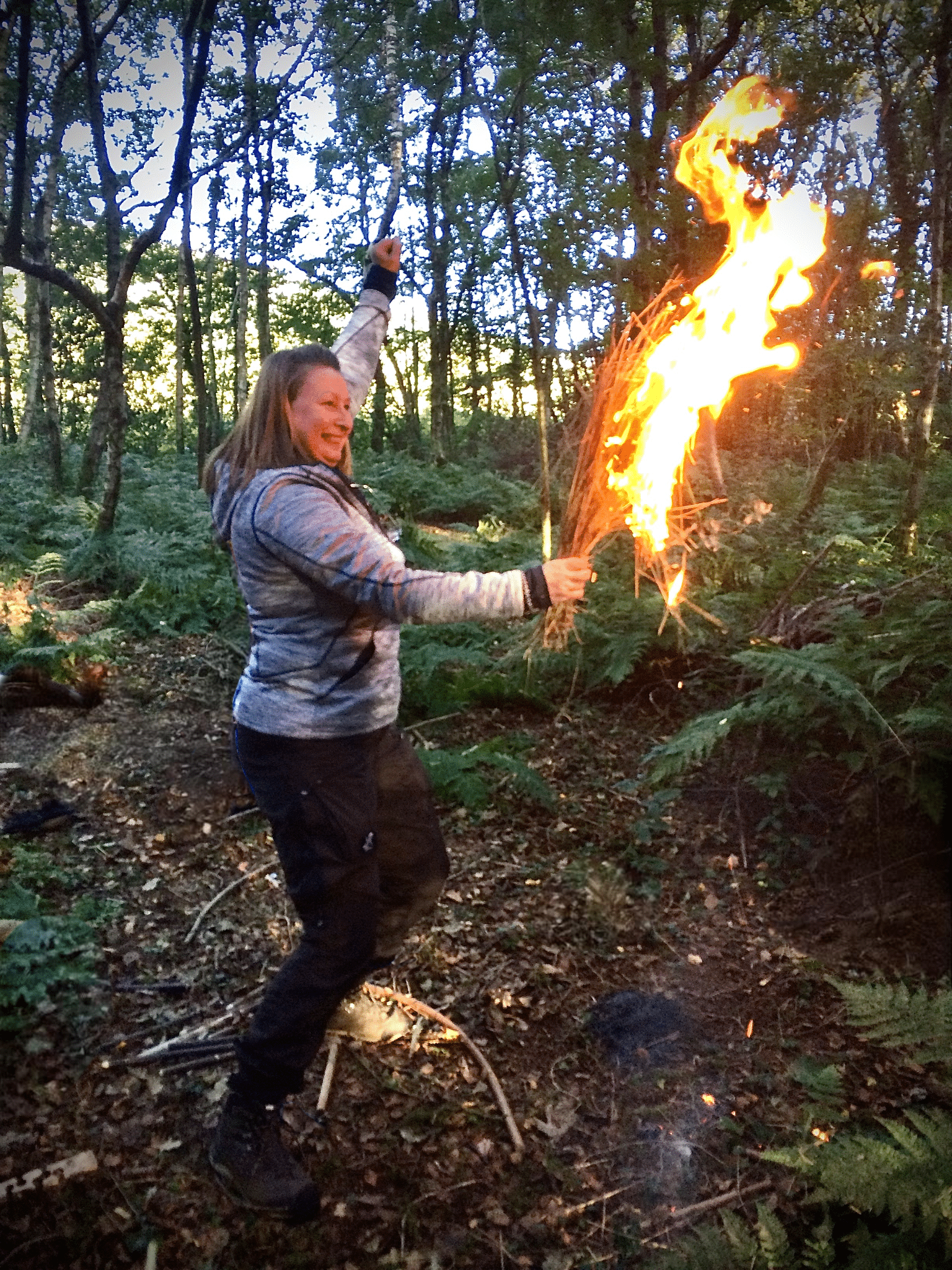Celebrating success with friction fire - a burning tinder bundle being held and a cheering hand. Large smile on women’s face. Weekend Bushcraft Course - Fire By Friction - Pippin & Gile - Bushcraft and Foraging - Courses - South London - Success