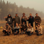 a group of people with a forbidding conifer block behind them in open grassland posed with a range of items including bows and axes