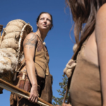 Theresa looking beyond the camera in full Stone Age clothing with a bow in hand and a pack frame on her back.